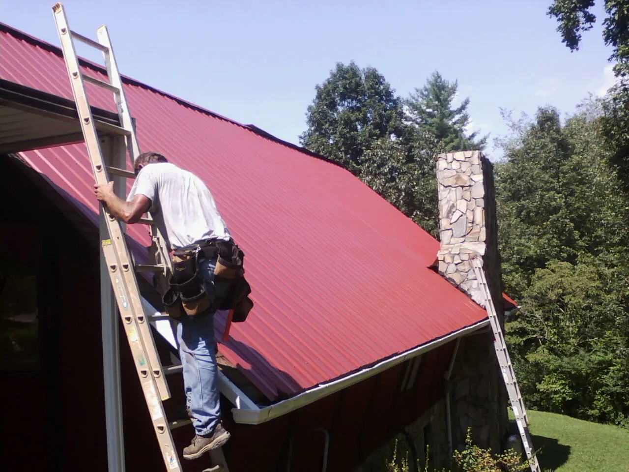 Roofer on a ladder beside a red metal roof and stone chimney — Soffit & Fascia Repair by Roofing Brothers in Westland